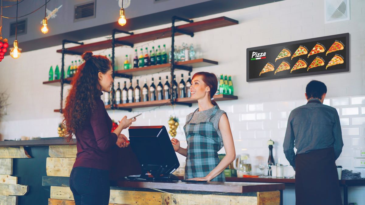 A woman paying a smiling cashier with the new PPDS Philips Tableaux zero-power series displaying digital signage.