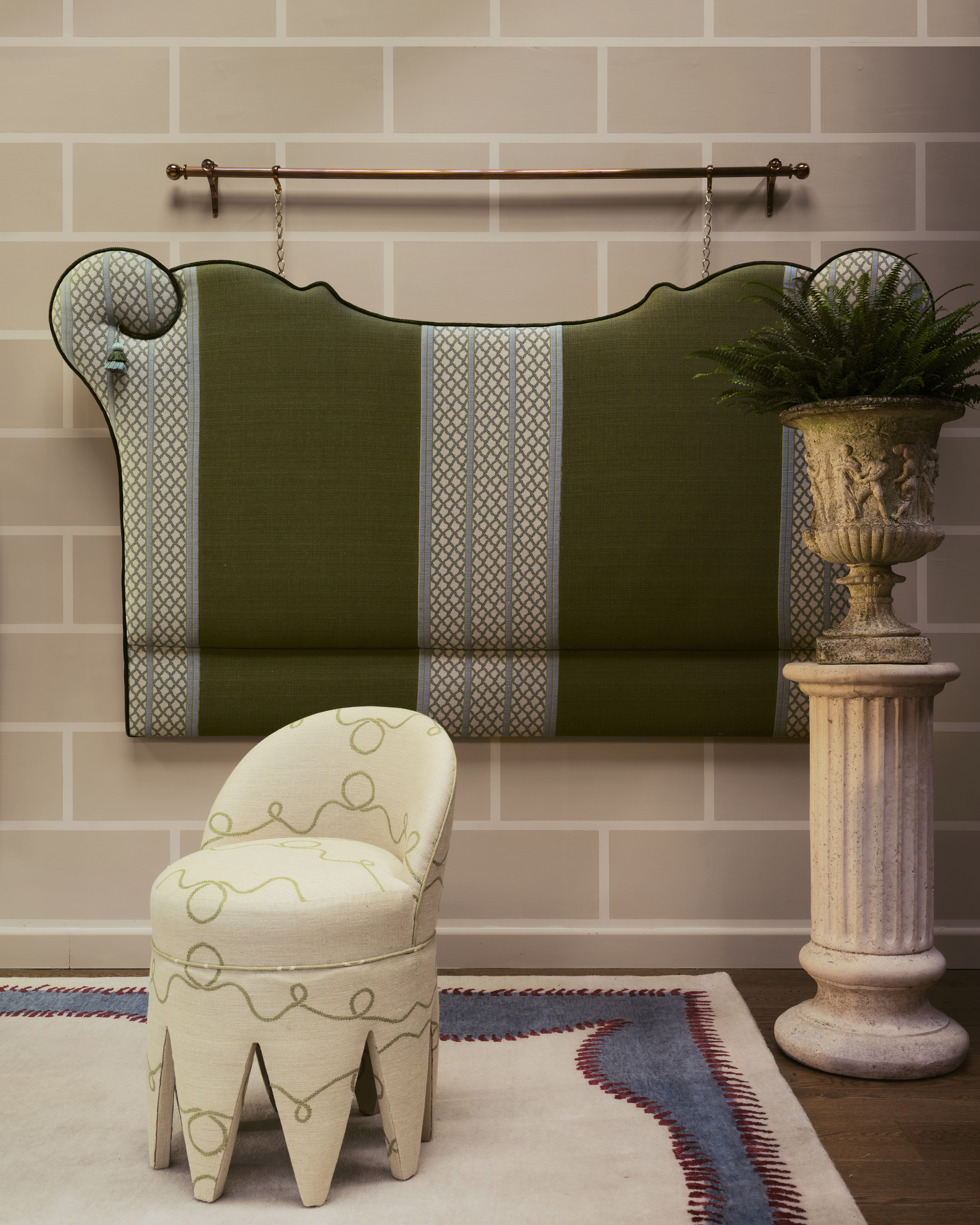 Image of a white stool seat with green lines on it on a white rug with blue and red trim. There is a beige wall with a blue and green patterned and striped headboard hanging fro a copper bar.