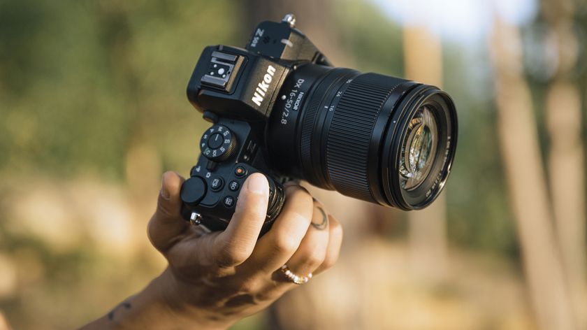 A person holds a Nikon mirrorless camera with a zoom lens, surrounded by a blurred, natural background