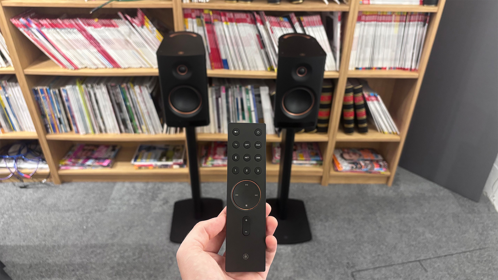 Yamaha NX-70A all-in-one system on stands in front of bookcase with remote control held in hand in foreground