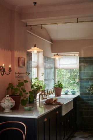 cottage style kitchen with pink walls, green backsplash, double sink with brass faucets, plants, scalloped pendants