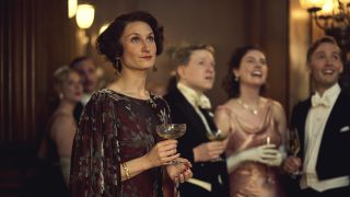 Bessie Carter in a dark floral dress holding a glass as Nancy Mitford in Outrageous