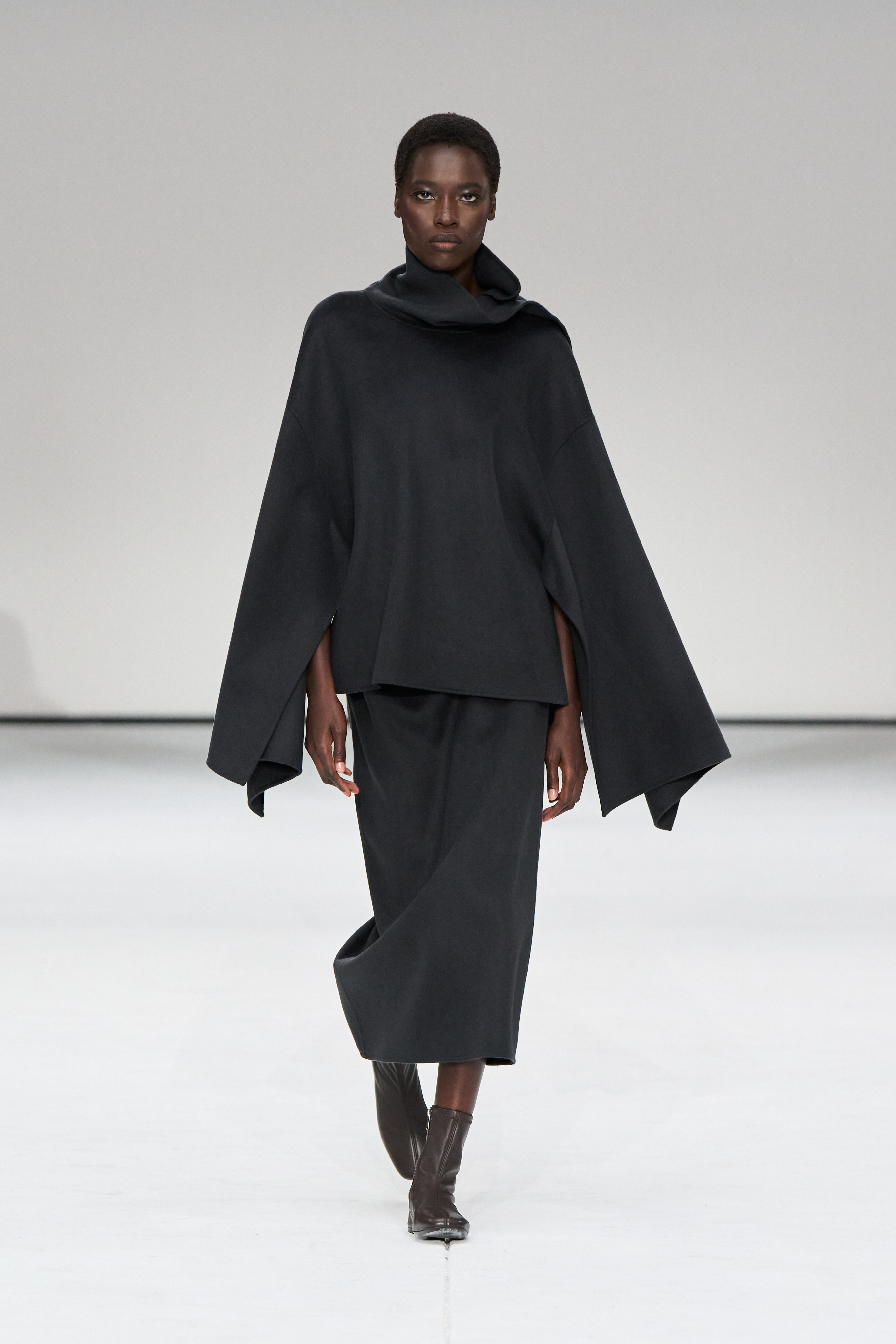 A woman wears a black poncho and black midi skirt with sock boots.