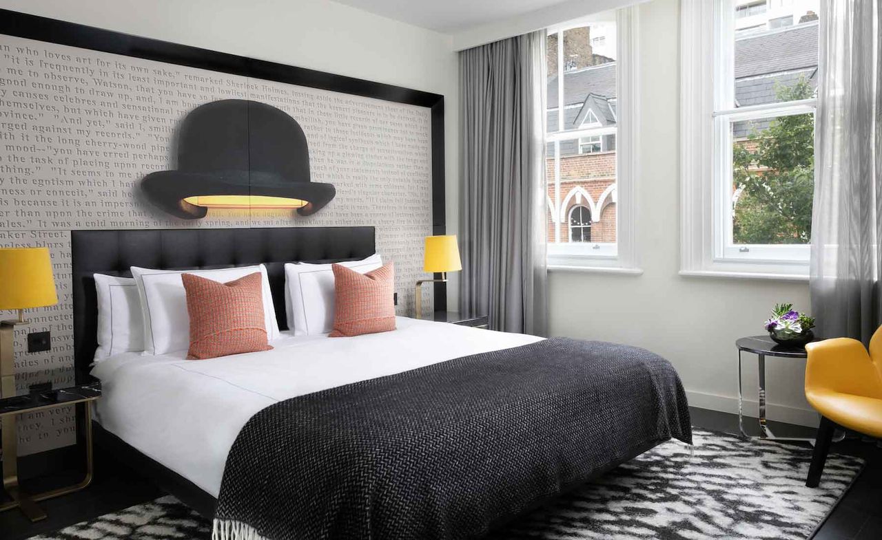 A room in the Hilton London Bankside hotel. A large bed is to the left. White linen, orange pillows, and a black blanket sit on top. A large bedframe in black and white covers almost the entire wall. There is writing over the whole headboard. Two large windows let in a lot of light. A yellow chair and lamps break up otherwise a very monochrome interior.