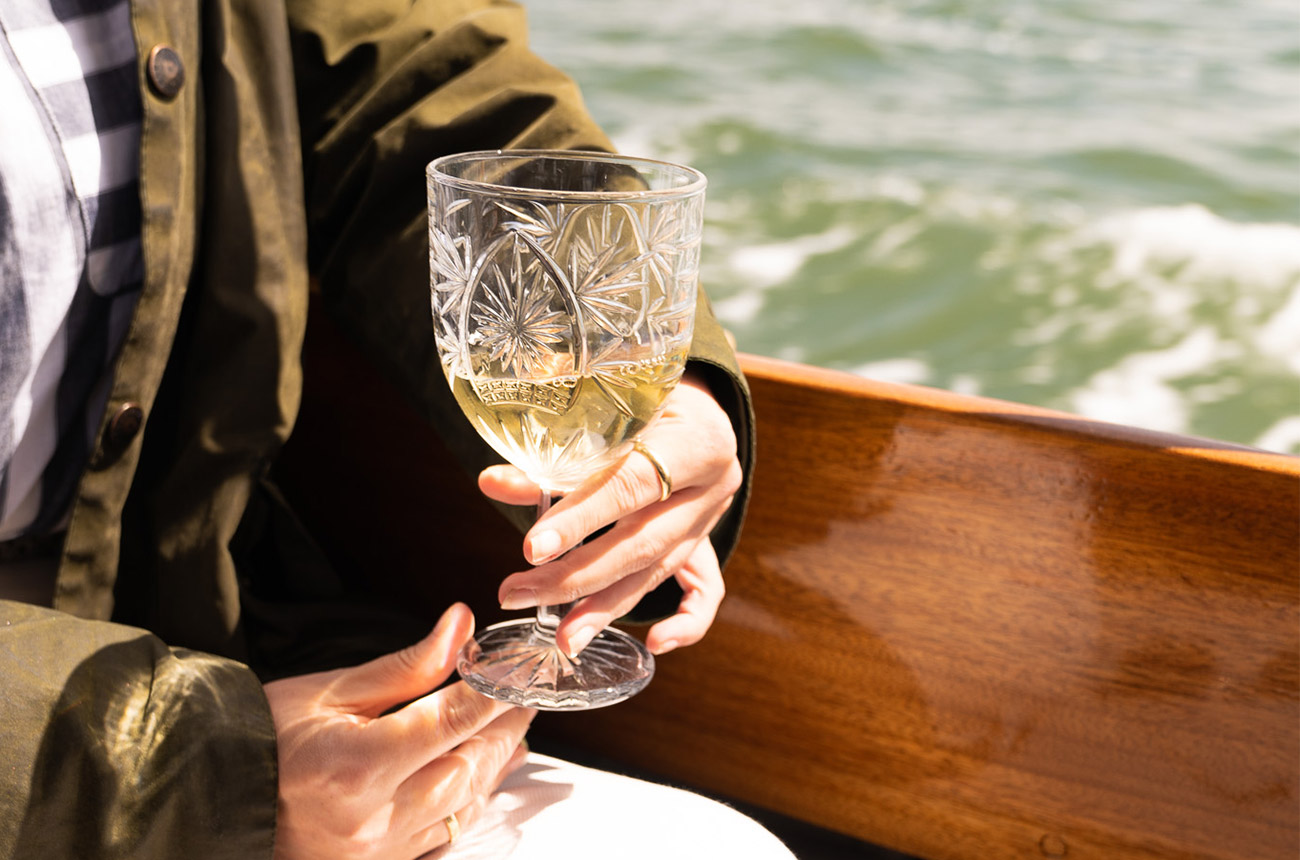 A close-up of someone on a boat holding a glass of white wine &amp;ndash; a hand holding the glass with water in the background