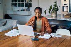 Person sits at dining room table with calculator, laptop and financial documents in front of her