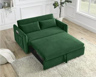 A green crushed velvet-style sofa bed folded out in a living room with a rug and lamp.