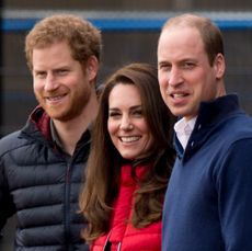 Prince Harry, Princess Kate and Prince William wearing coats standing next to each other and smiling at the camera