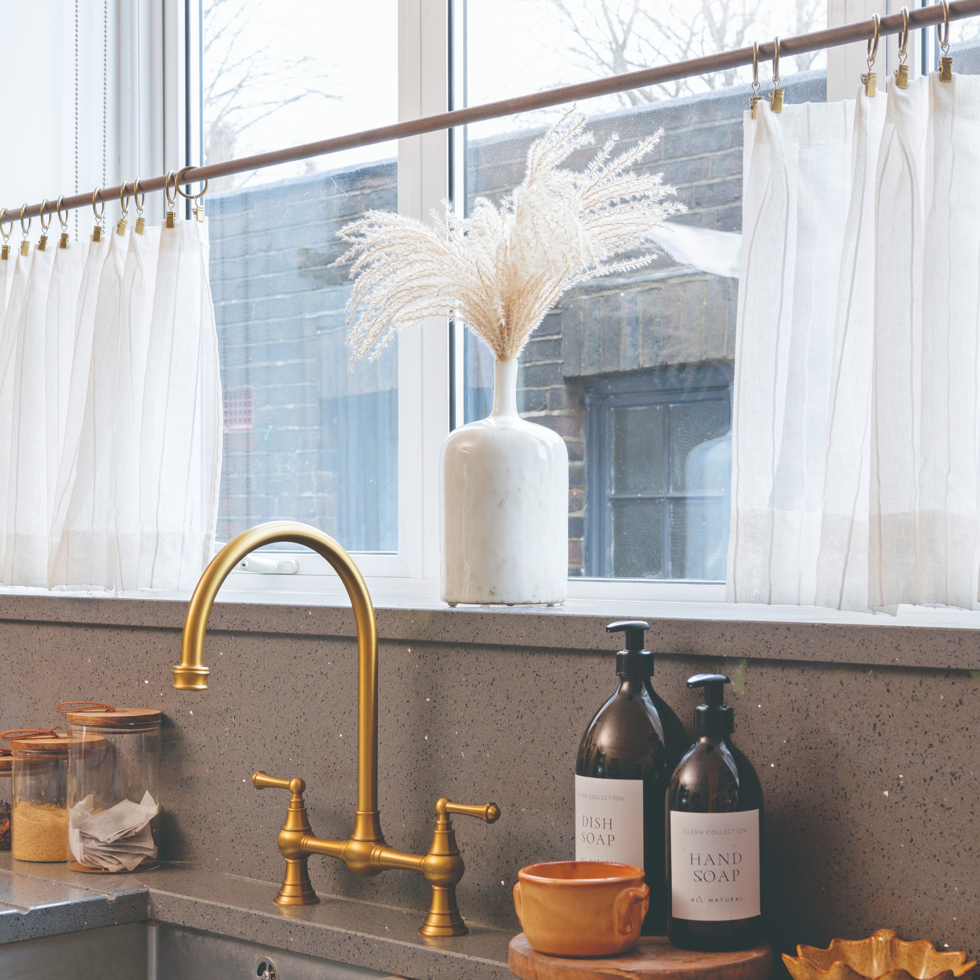 A kitchen sink with a brass tap with semi-sheer white cafe curtains on the window above