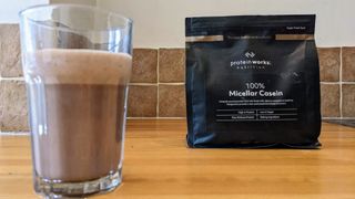Protein Works 100% Micellar Casein mixed in glass with packet in background
