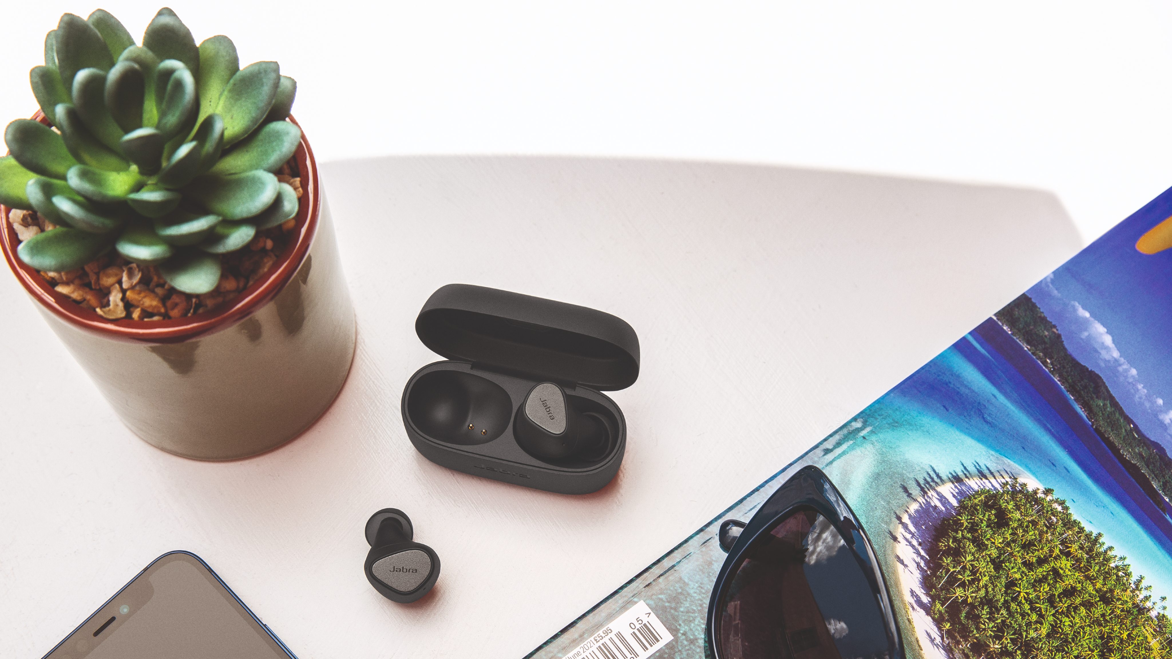 Jabra Elite 4 på et bord ved siden af en grøn plante og et rejsemagasin
