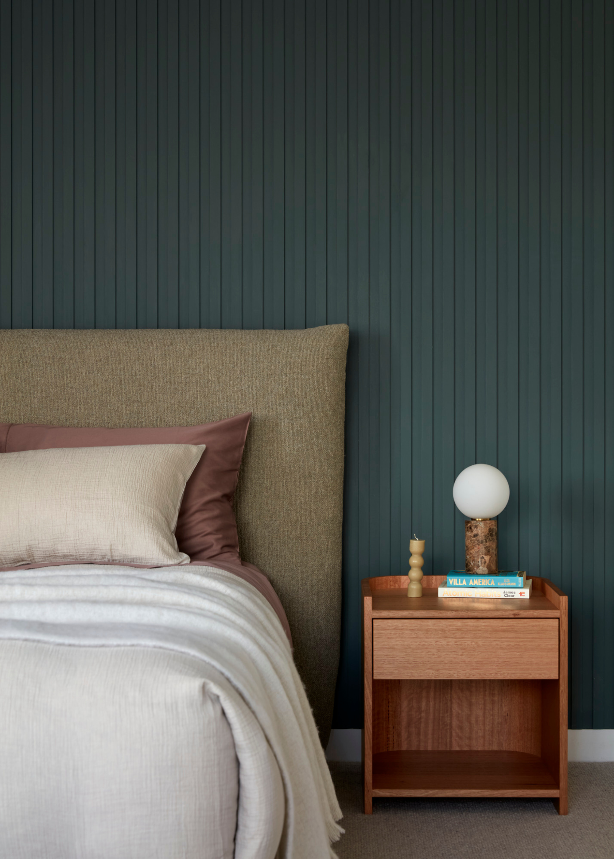 A bedroom with green paneled walls, a wood side table with books and a lamp, and a bed with a brown headboard and pillows