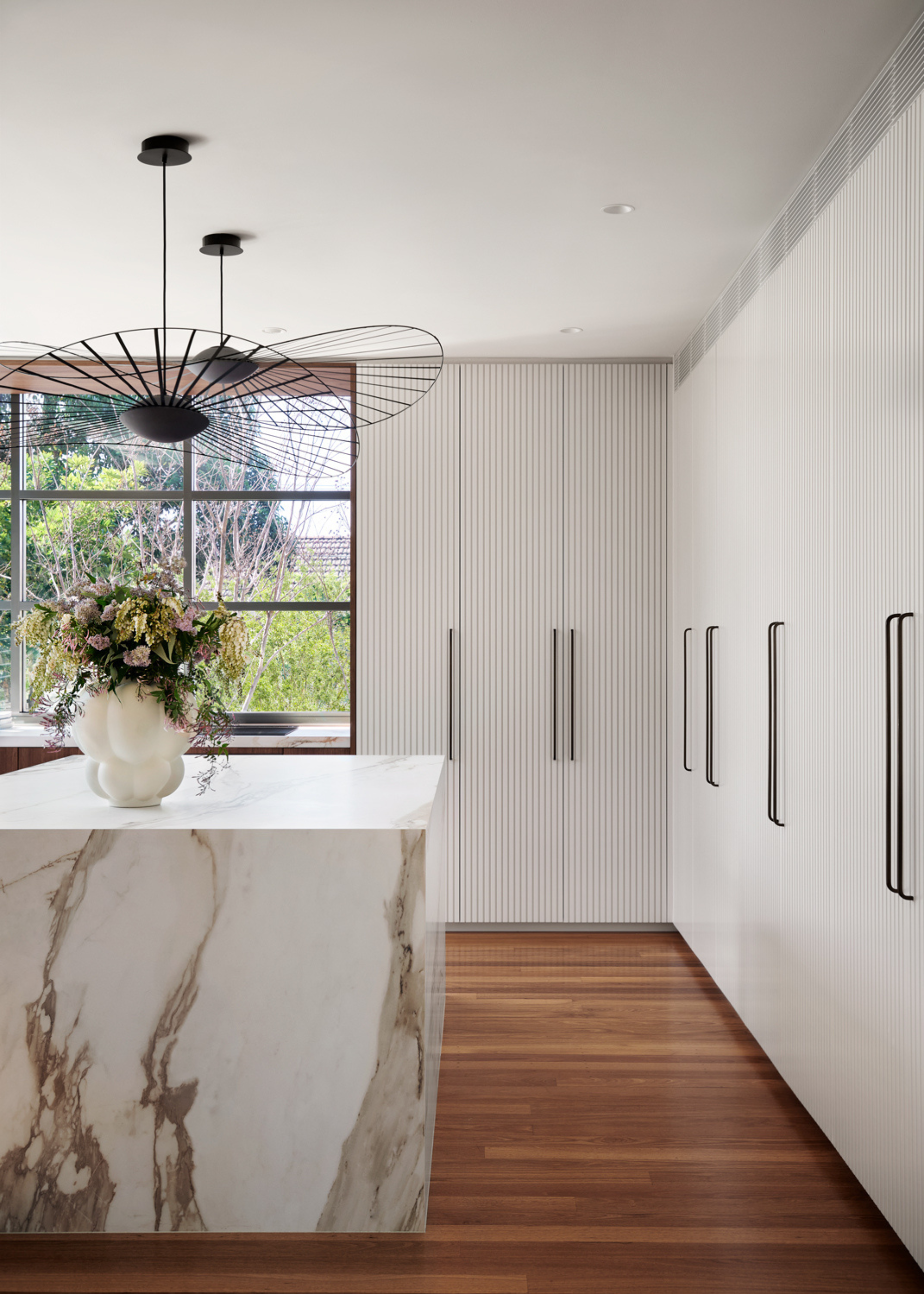 An all-white modern kitchen with floor-to-ceiling fluted cabinetry and long black modern handles, with a white and grey marble waterfall island and a vase of flowers styled on top