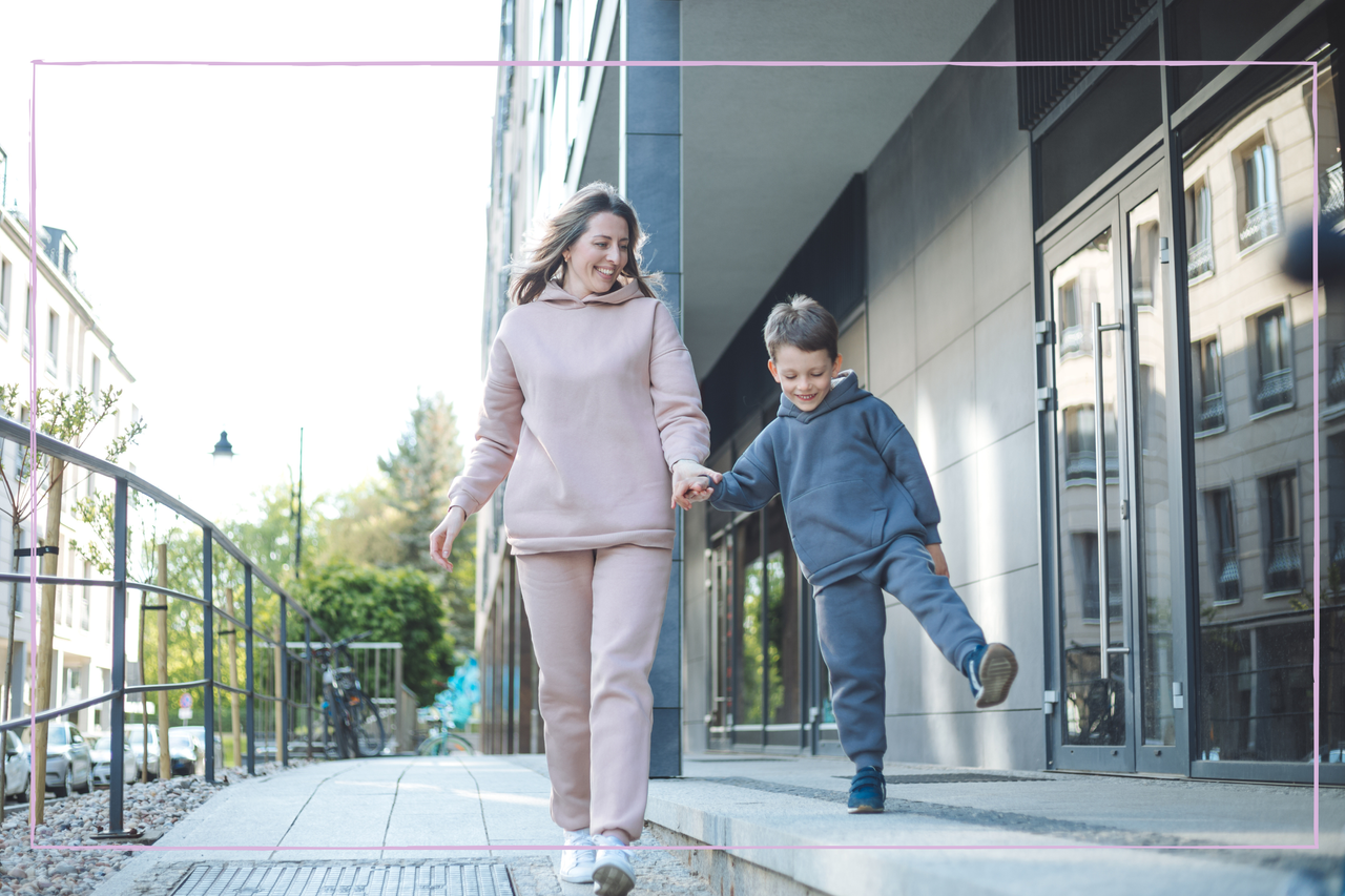 Slim middle-aged woman with brown hair in beige sportswear walking on sidewalk with her little boy with short brown hair in blue tracksuit holding hands and smiling