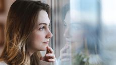 Thoughtful young woman looking out the window, her reflection visible on the glass