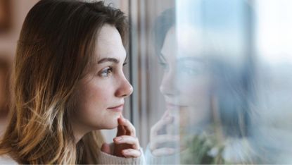 Thoughtful young woman looking out the window, her reflection visible on the glass