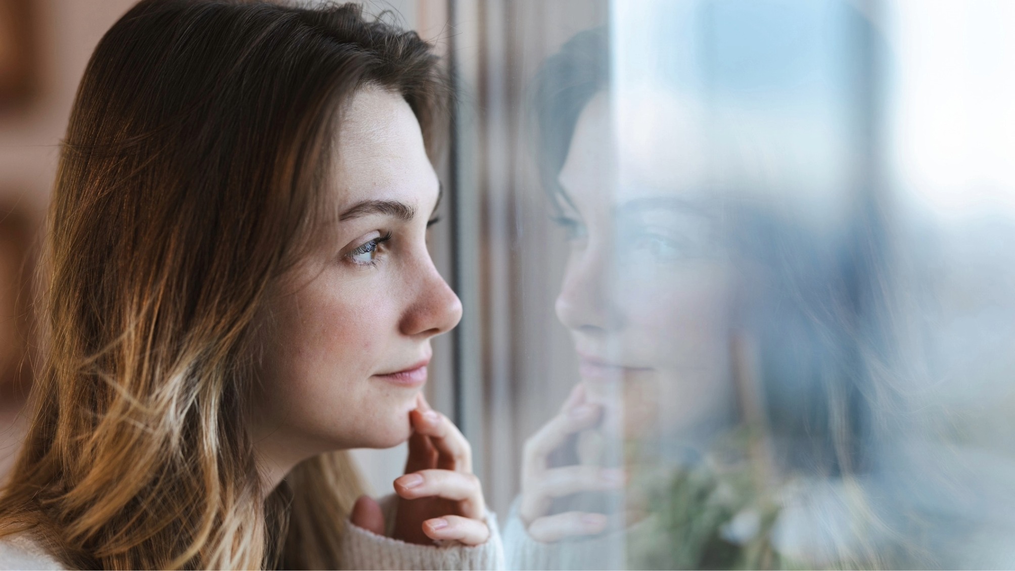Thoughtful young woman looking out the window, her reflection visible on the glass