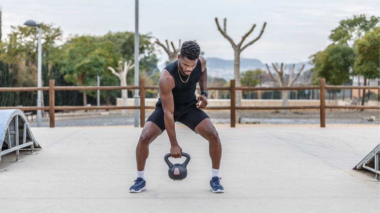 A man holds a kettlebell in an outdoor setting. He is squatting slightly and gripping the kettlebell in one hand between his legs. Behind him we see trimmed trees and a wooden fence.