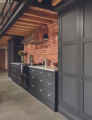 Dark green-gray painted kitchen cabinets in a characterful room with exposed brick walls and stone flooring.