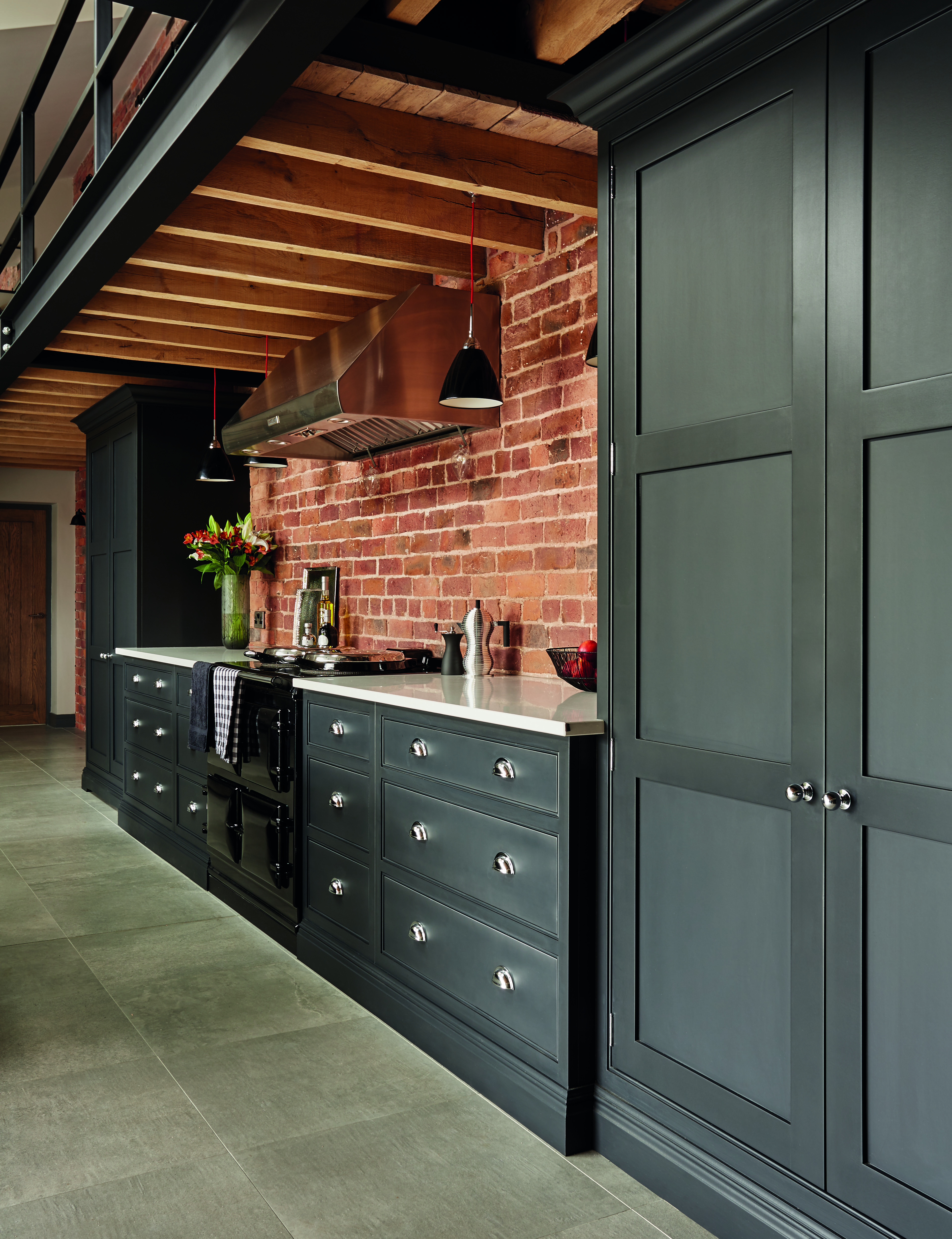 Dark green-gray painted kitchen cabinets in a characterful room with exposed brick walls and stone flooring.