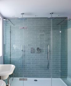 A blue tiled shower in a bright bathroom with a window, white matching sinks, purple glasses above with a yellow bottle of hand soap, and a silver radiator with two white towels hanging up