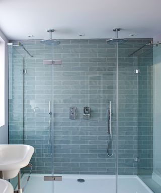 A blue tiled shower in a bright bathroom with a window, white matching sinks, purple glasses above with a yellow bottle of hand soap, and a silver radiator with two white towels hanging up