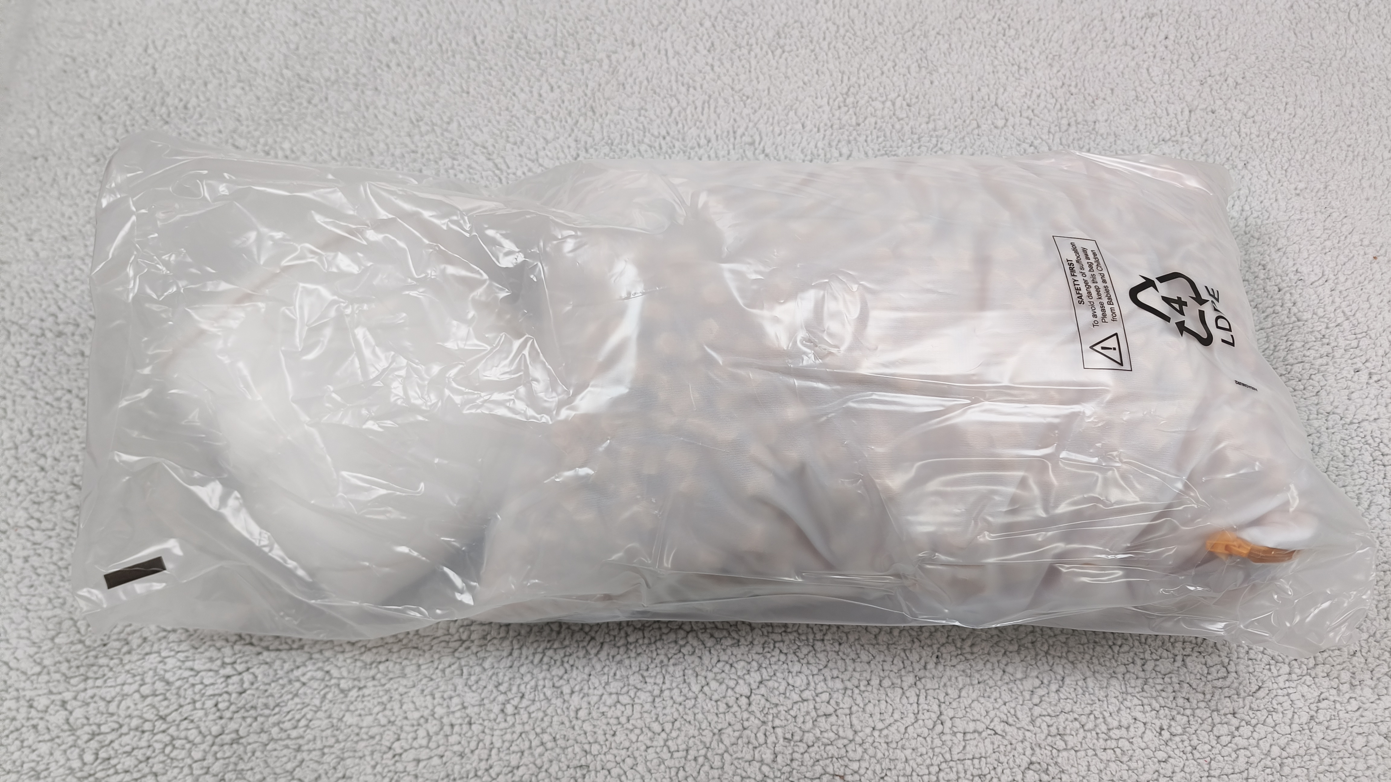 A transparent plastic shipping bag containing a folded white pillowcase and a mesh bag of orange foam clusters, resting on a gray textured rug.