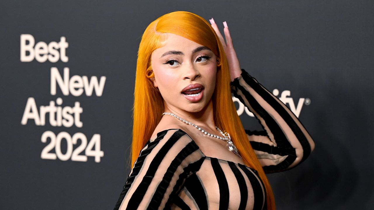 Ice Spice stands in front of a step and repeat while wearing a striped semi sheer bodysuit