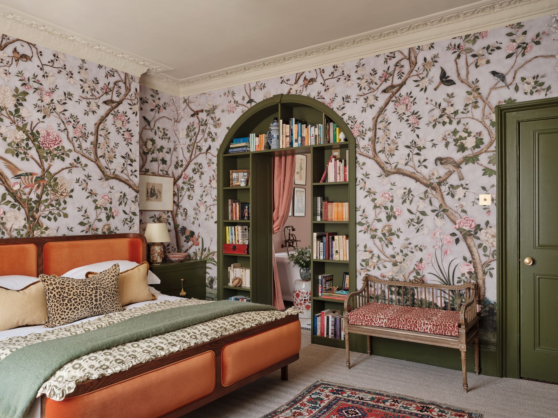 A bedroom with a botanical wallpaper, green trim, a built-in bookcase around the door, and a large orange bed