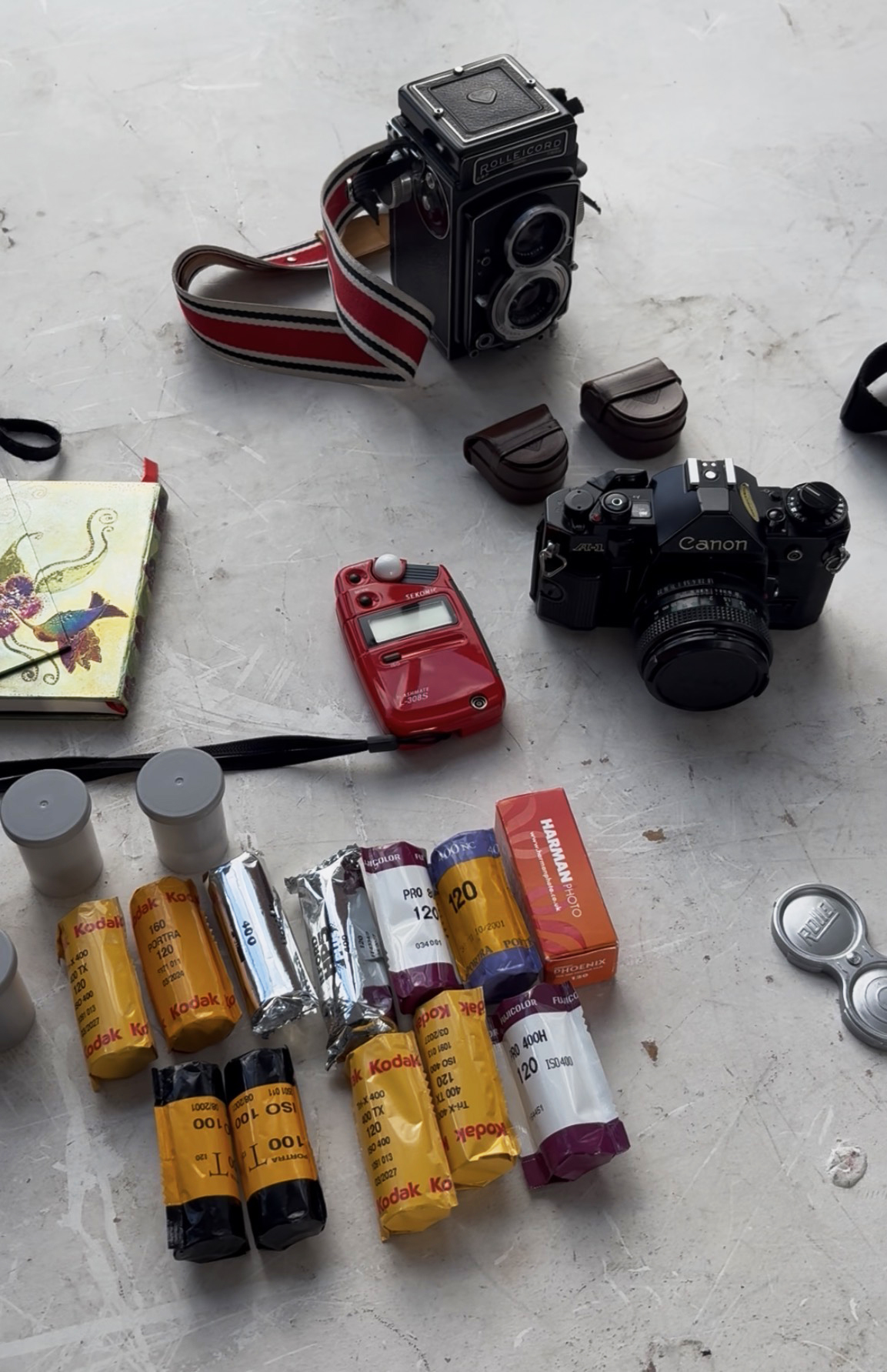 Two film cameras, a light meter, and various rolls of film scattered on a table, alongside a colorful notebook