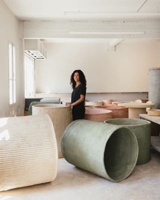 A young woman dressed in black clothes stands in a sun-lit studio surrounded by large cement vases in neutral colors.