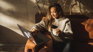 Woman using a credit card to shop on her laptop sat on the sofa wearing a sweater.