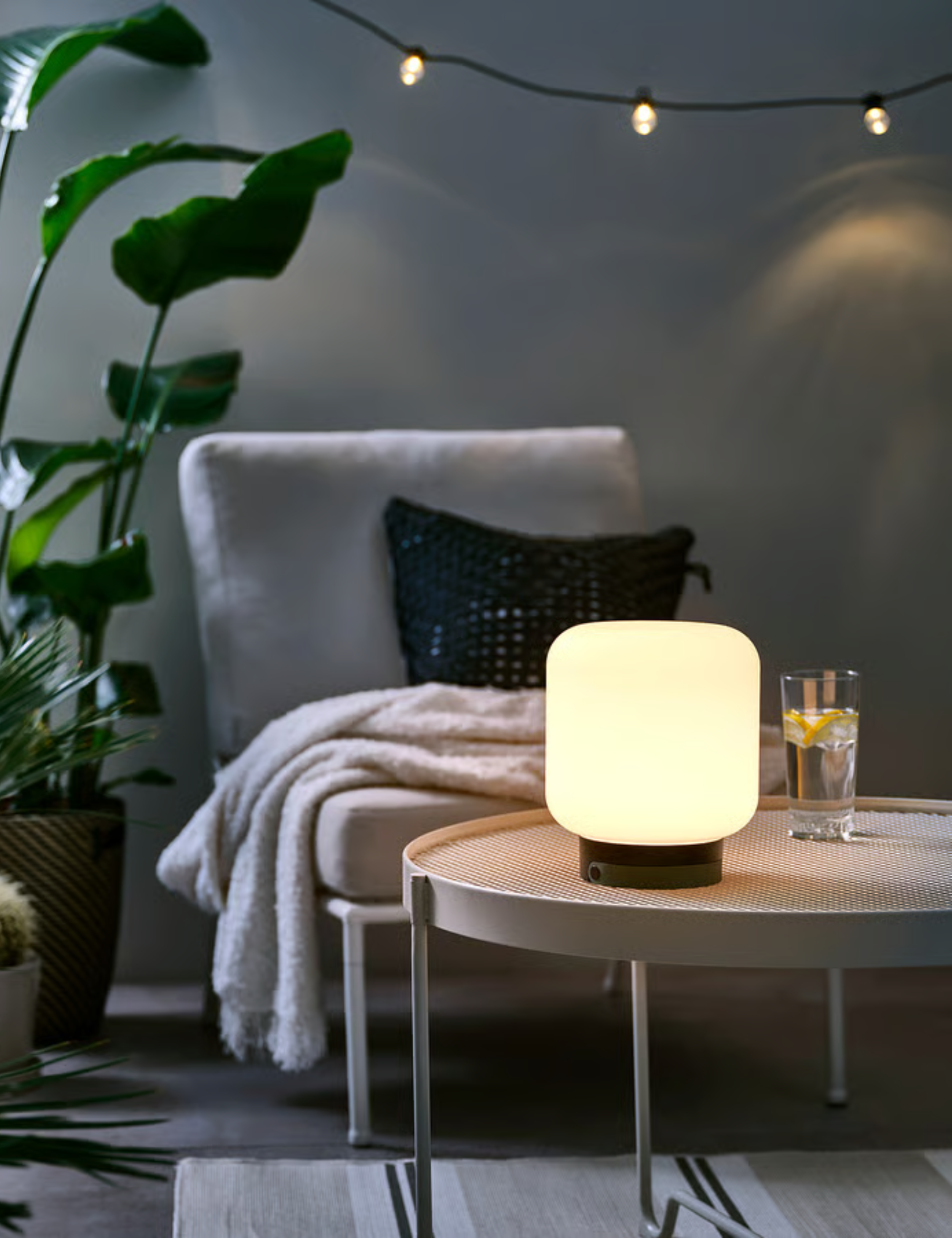 IKEA outdoor L&amp;Auml;NSPORT lamp pictured atop of a wooden patio table alongside a lounge chair and various potted plants.