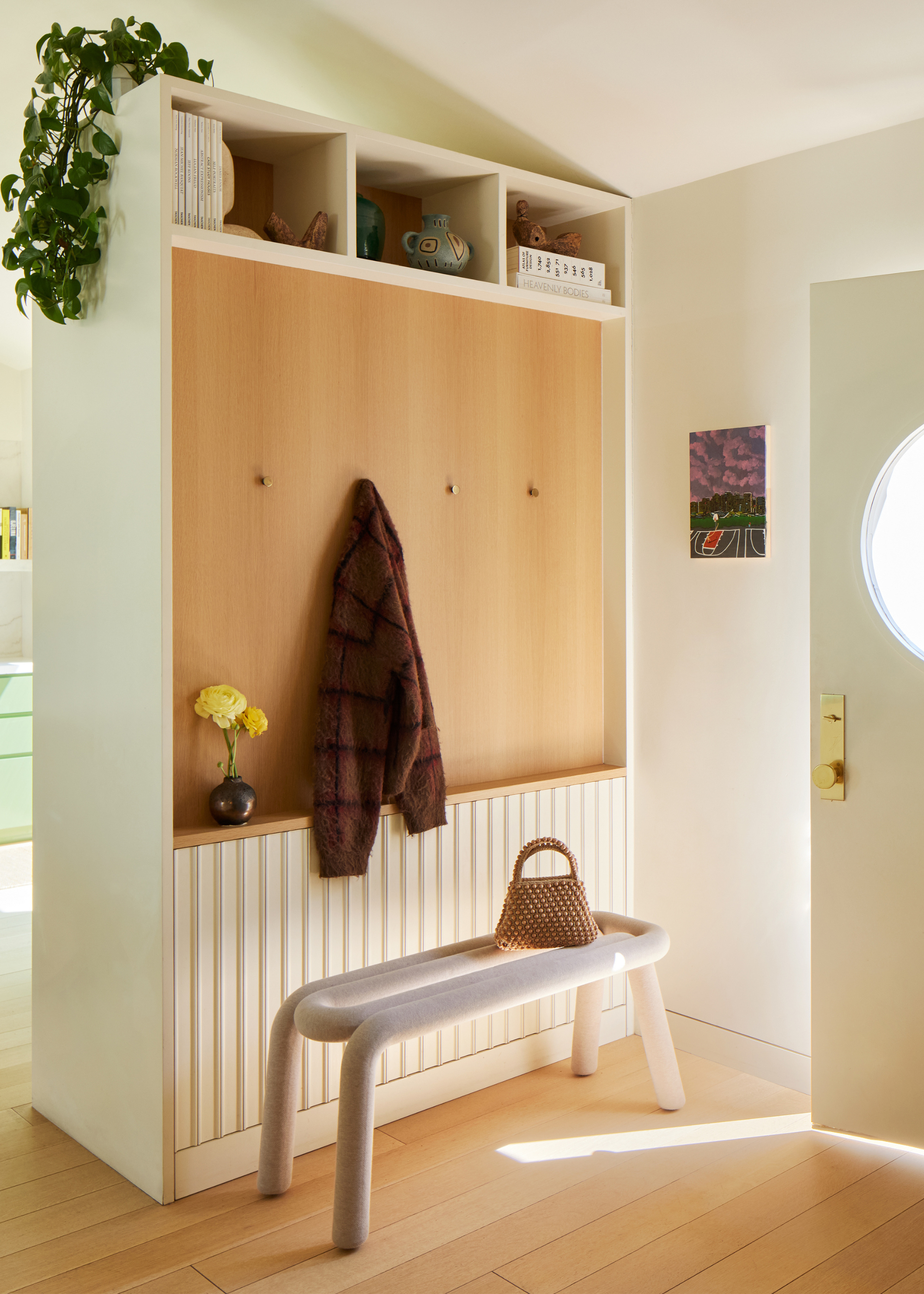 An entryway with a hooks for coats, a vase of flowers, a white tubular bench, a shelf for books, vases, and decor, alongside a trailing plant