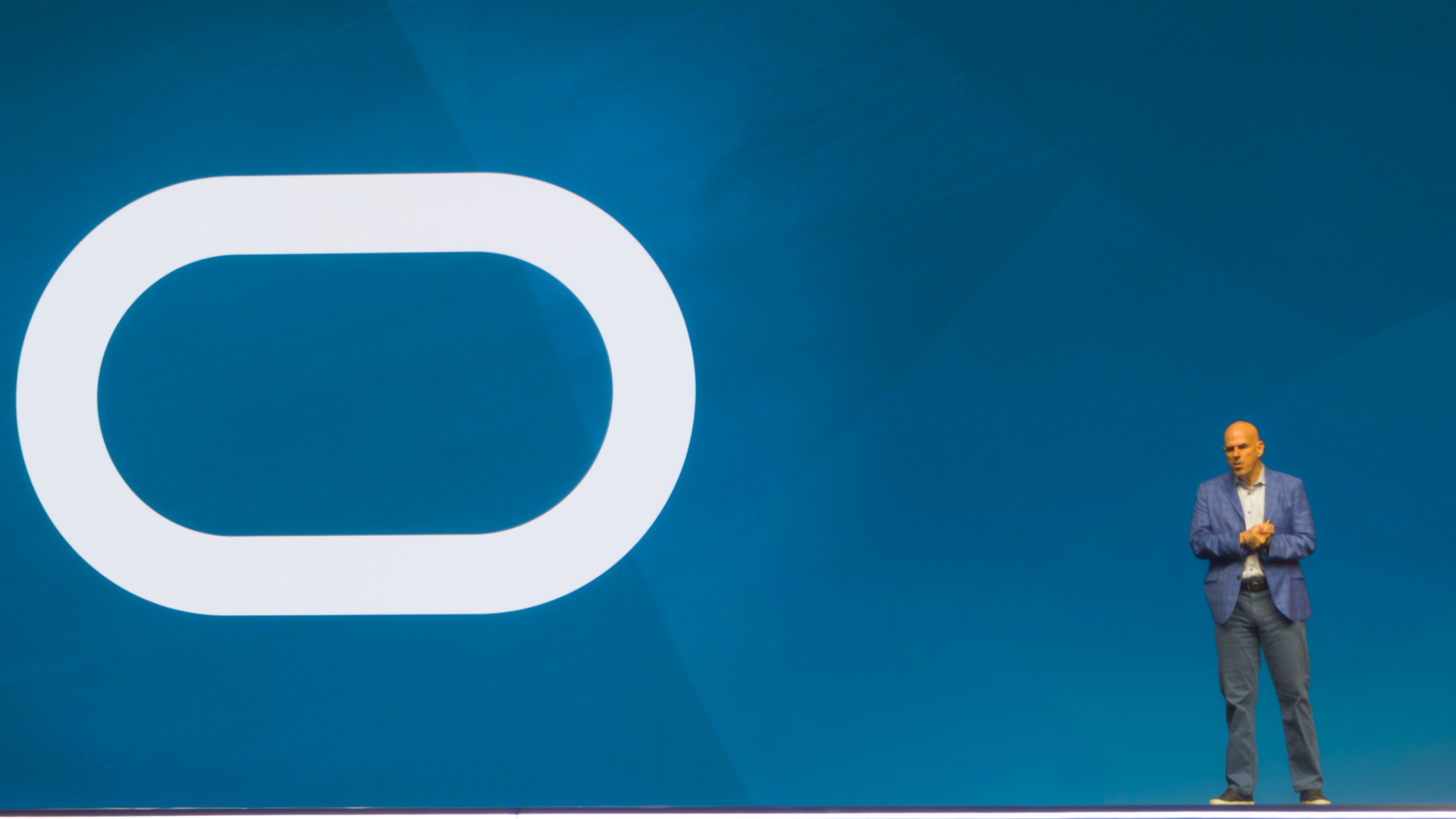 A photo of Mike Sicilia, CEO at Oracle Corporation, onstage at Oracle AI World Tour London 2026. Sicilia is stood against a blue background, with the large blue 'O' of the Oracle logo to his left. 