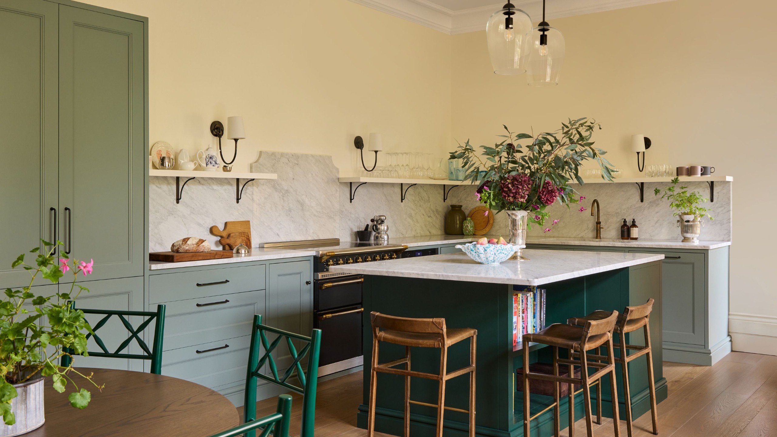A large kitchen in a traditional country home with off-white walls, sage green cabinets, and a dark green island. White marble countertops and backsplash, open shelving, and antiqued brass wall lights.
