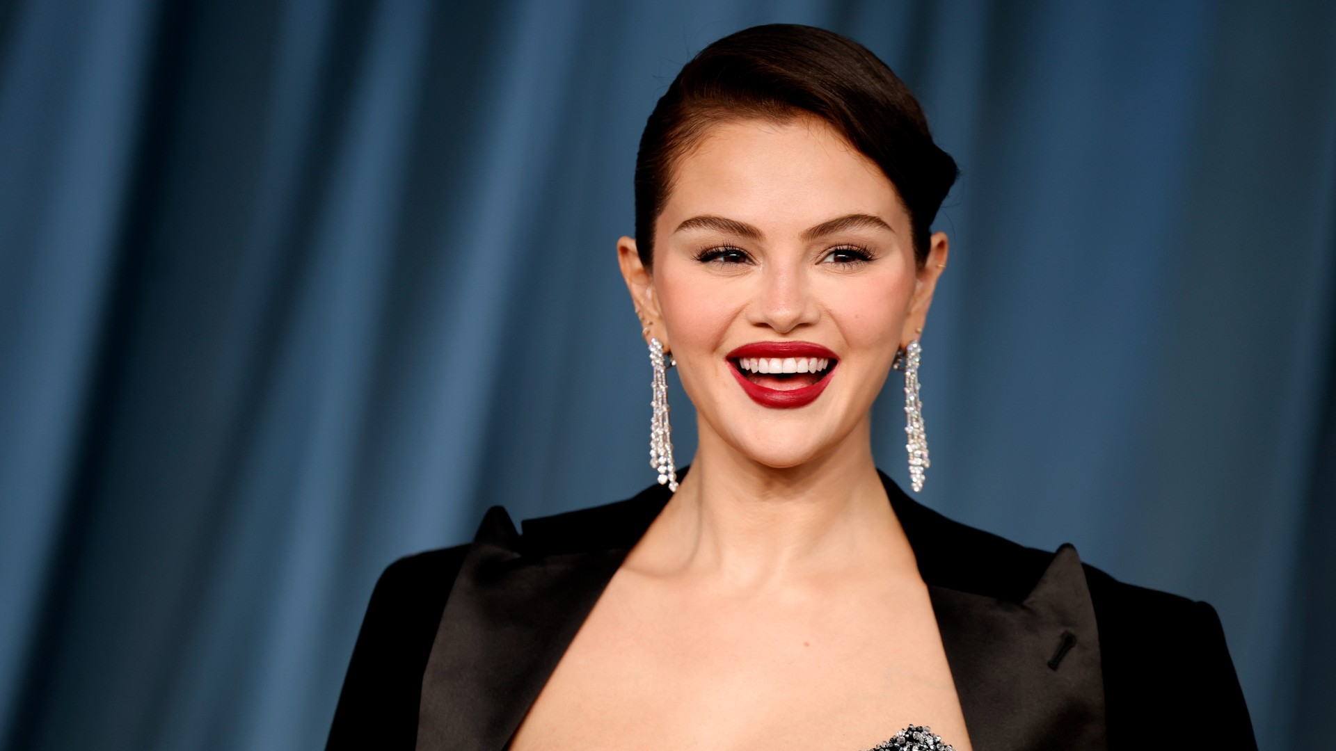 a woman with dark hair wearing a black blazer and an updo posing for paparazzi in front of a blue background