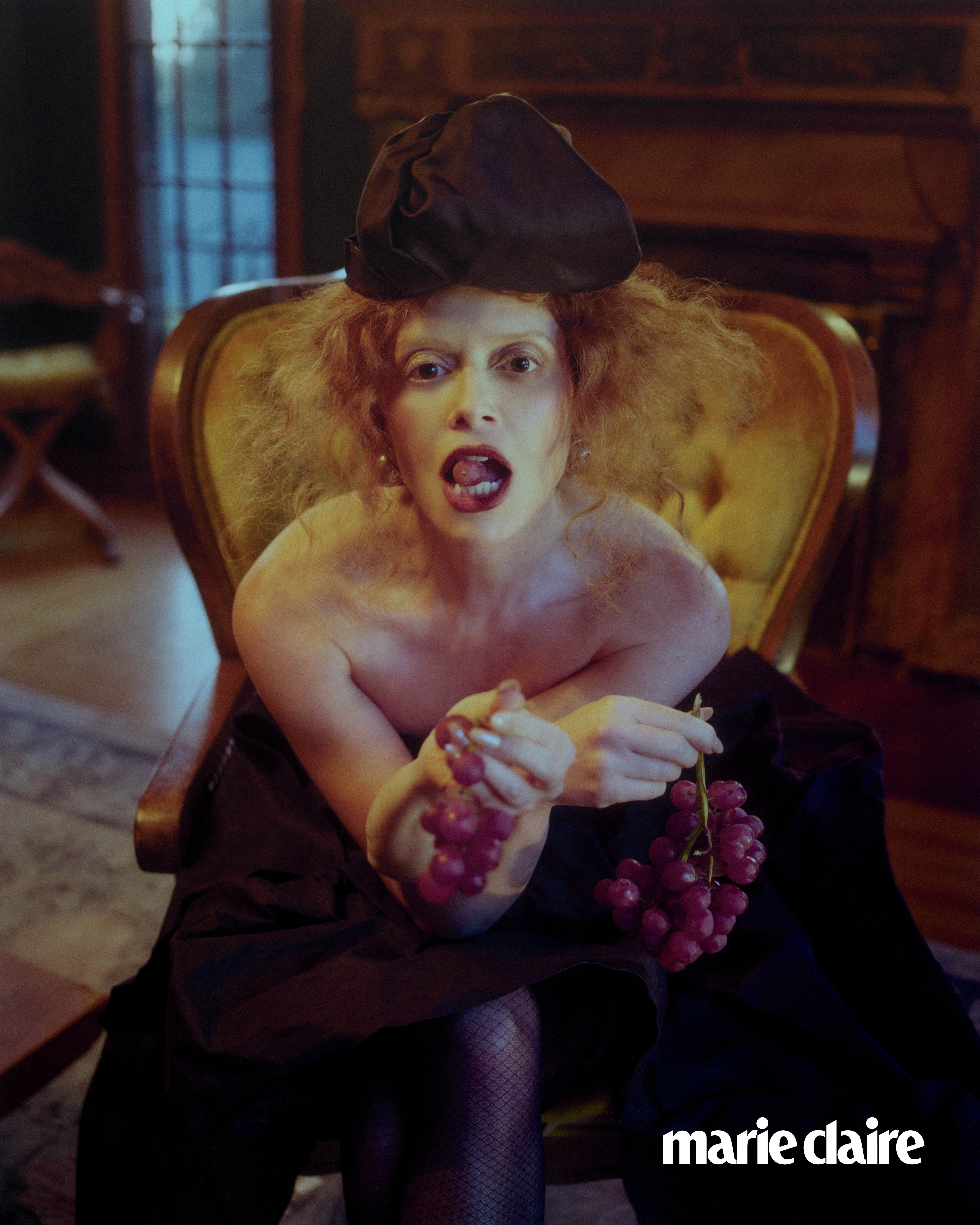Natasha Lyonne leaning forward with fruit in her mouth wearing a beret in a shoot for Marie Claire