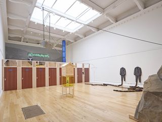 An exhibition space featuring artwork including two suits on hangers on a wall, a large bird feeder and house, multiple doors next to each other, an illuminated sign saying Johnnys.