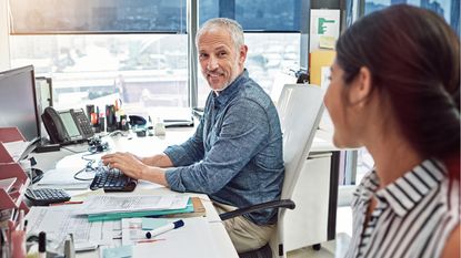 An older man working on a computer at a desk in an office smiles at a co-worker.