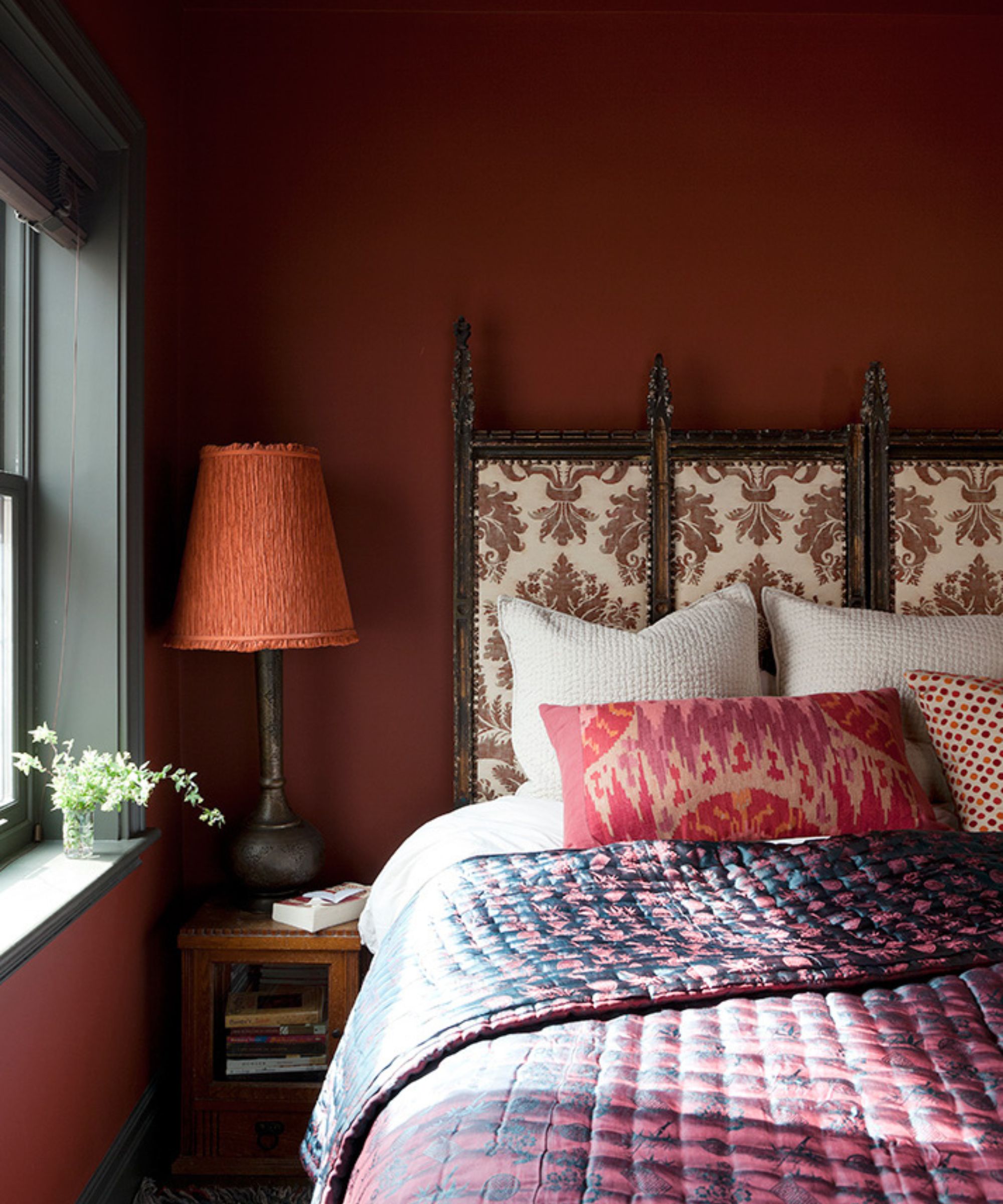 A cozy bedroom with dark red walls, a patterned upholstered headboard, layered bedding with a dark blue and raspberry red quilt and red cushions. A nightstand with a terracotta lampshade and black window trim.