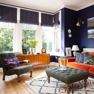 blue living room with orange sofa, sage green armchair and footstool, patterned rug, mid-century sideboard, graphic blinds, wall light, bold graphic cushions