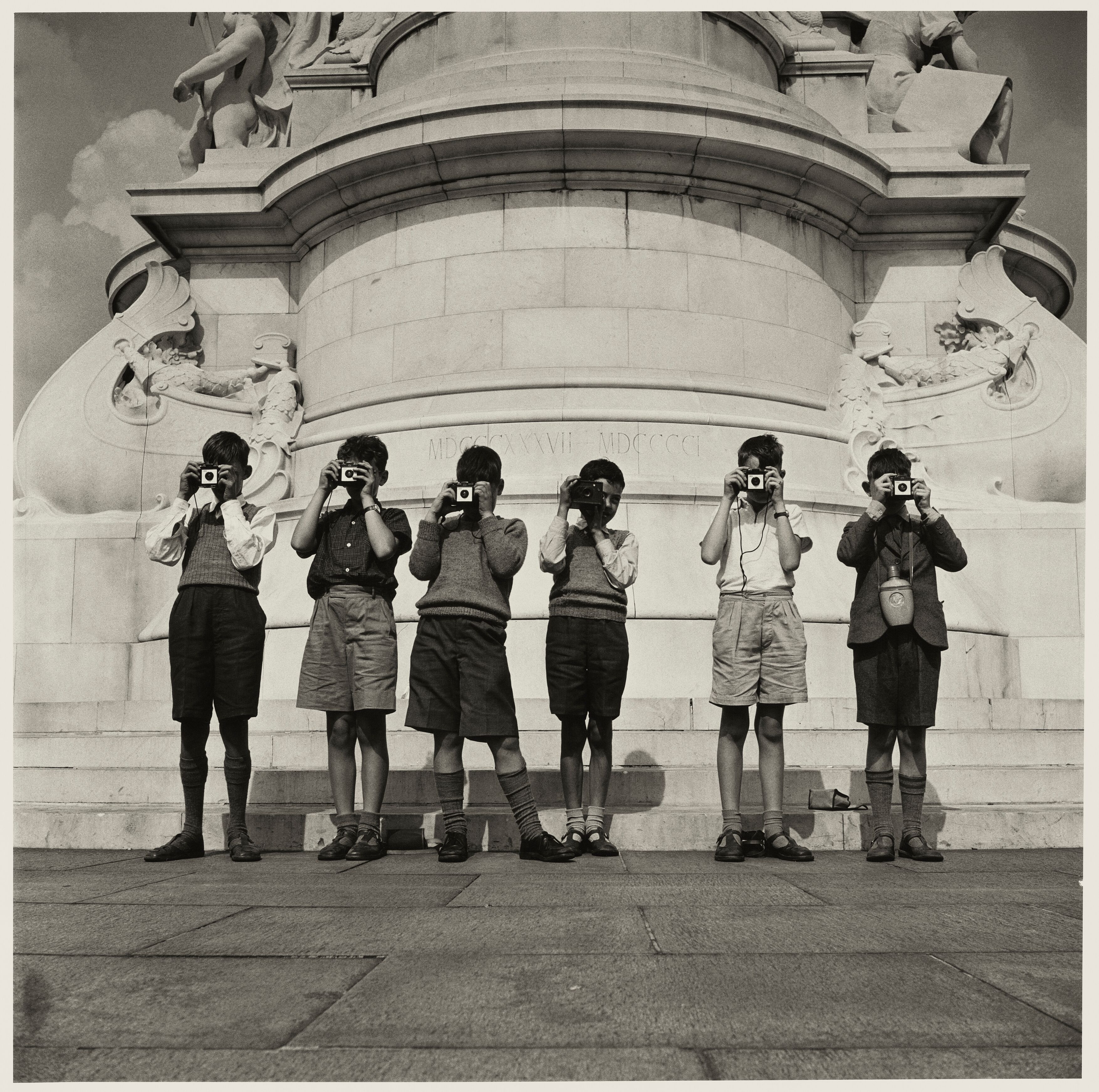 Six young boys stand in a line against the base of a stone monument, each holding a small camera up to their eyes to take a picture.