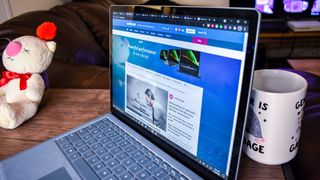 En Surface Laptop 4 står öppnad på ett skrivbord bredvid en teddybjörn och en kaffekopp. TechRadar-hemsidan visas i webbläsaren.