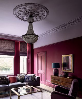 Large living room with two-toned purple painted ceiling and walls, large glass beaded chandelier, wooden flooring, purple rug, purple armchair and sofa, glass square coffee table, wooden sideboard