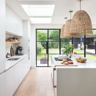 Metal framed glazing including patio doors and fixed full height windows in contemporary kitchen with white cabinets and rattan pendant lights over the kitchen island