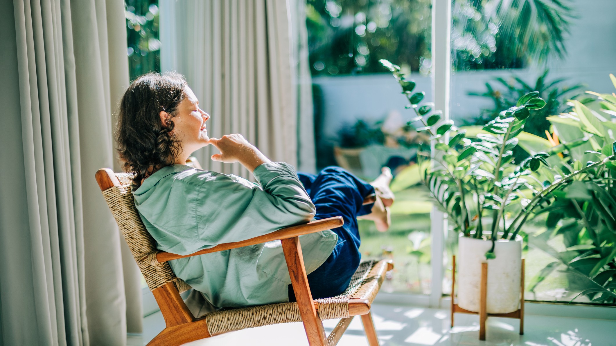 A woman lounges on a wicker chair in a sunny window.