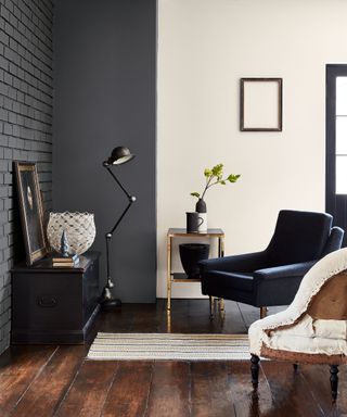 Living space with graphite painted brick wall on ceiling, cream painted wall to the right, dark wooden flooring, lounge chair in blue and lounge chair upholstered in cream, low black table with painting and decorative bowl, black floor lamp, metal side table with vases, small patterned striped rug
