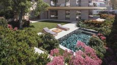 A large garden with pink flowers and layered shrubs leading to a pool by a set of sun loungers