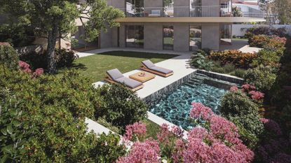 A large garden with pink flowers and layered shrubs leading to a pool by a set of sun loungers